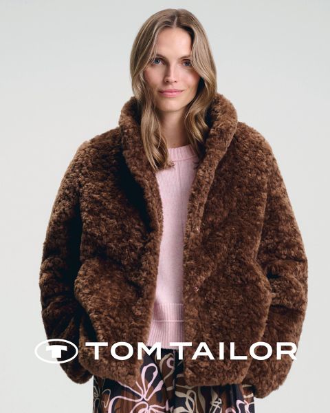 Eine lächelnde Frau in einem kastanienbraunen Pullover und ein Mann in einem marineblauen Pullover stehen Seite an Seite an einem Strand. Beide tragen Jeans. Der Himmel ist klar und blau. Das Logo "Tom Tailor" erscheint am unteren Rand.