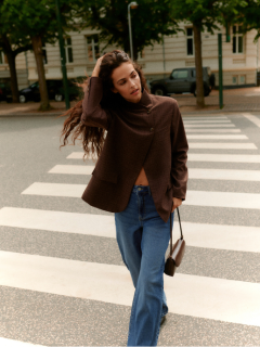 Eine Frau mit lockigem Haar steht an einer gekachelten Wand. Sie trägt eine hellgraue, wuschelige Strickjacke, schwarze, hoch sitzende Ledershorts und eine schwarze Strumpfhose, die Hände in den Taschen und einen selbstbewussten Gesichtsausdruck.