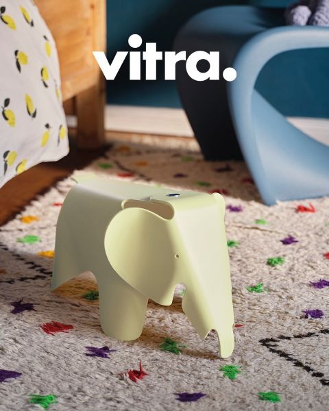 Ein modernes Wohnzimmer verfügt über ein rosa-beige gestreiftes Sofa, einen weißen Stuhl mit Holzbeinen und einen runden Couchtisch aus Holz. Auf dem Tisch steht eine Vase mit bunten Blumen, darüber steht „vitra.“ an der Wand. Der Boden ist mit einem weichen Teppich bedeckt.