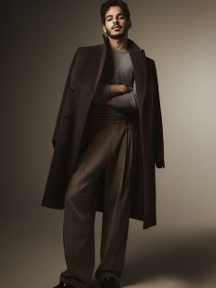 Ein Mann steht selbstbewusst da, trägt einen langen, dunklen Mantel über einem grauen Pullover und eine weitbeinige braune Hose, die Arme verschränkt. Der Hintergrund ist schlicht und sanft beleuchtet, was eine stilvolle und anspruchsvolle Atmosphäre schafft.