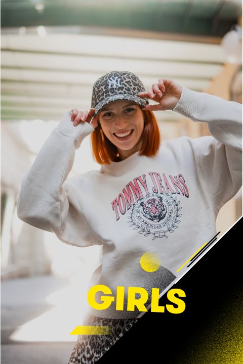 Eine lächelnde Person trägt ein graues „Tommy Jeans“-Sweatshirt und eine Leoparden-Cap. Die Cap sitzt mit beiden Händen zurecht. Im Hintergrund ist eine sonnige Außenkulisse zu sehen. Das Bild ist mit dem gelben Text „Girls“ überlagert.
