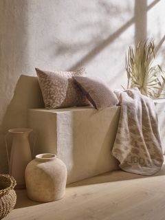 Ein minimalistischer Raum verfügt über eine beige Steinbank mit zwei strukturierten braunen und beigen Kissen, einer gemusterten Überwurfdecke und getrockneten Gräsern. Ein geflochtener Korb, eine rustikale Keramikvase und ein hoher Krug schmücken den Boden unter sanftem, natürlichem Licht.