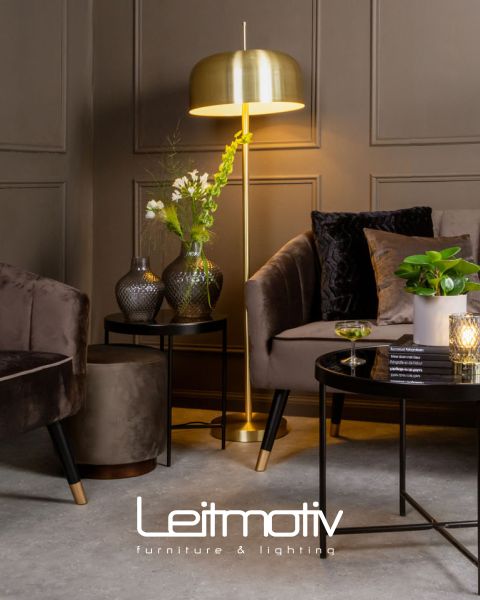 Ein stilvolles Wohnzimmer mit einer goldenen Stehlampe, dunklen Samtmöbeln und dekorativen Vasen auf Beistelltischen. Auf einem Couchtisch stehen ein Glas und eine Kerze. Die getäfelte Wand bildet einen eleganten Hintergrund. Unten befindet sich das Logo „Leitmotiv Möbel & Beleuchtung“.