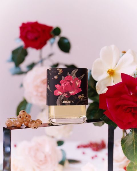Parfümflakon mit floralem Design und der Aufschrift „Liberty“ auf einer Glasoberfläche. Umgeben von roten und weißen Rosen, grünen Blättern, roten Johannisbeeren und einem unscharfen Hintergrund mit weiteren Blumen.