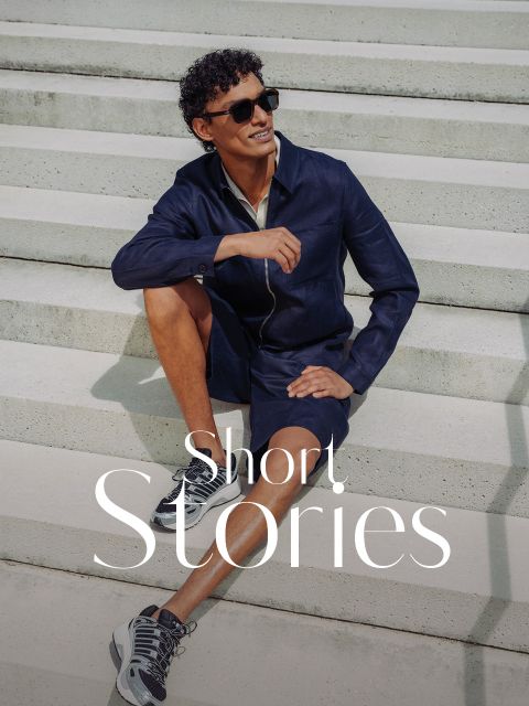 Ein Mann mit Sonnenbrille, marineblauer Jacke und kurzen Hosen sitzt lächelnd auf einer Freitreppe. Die Worte "Short. Stories" erscheinen am unteren Rand.