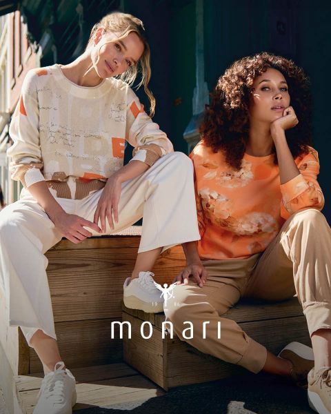 Eine lächelnde Frau, die einen beigen Mantel, ein gestreiftes Hemd, blaue Jeans und einen hellen Schal trägt, steht im Freien vor einem hohen Gebäude. Das Monari-Logo ist am unteren Rand des Bildes zu sehen.