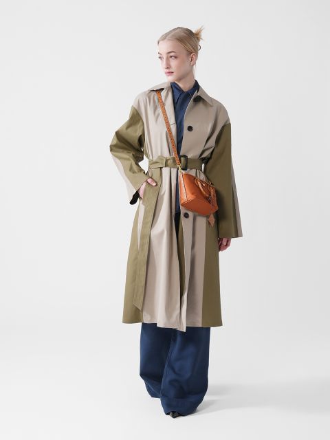 Eine Person steht in einem Studio. Sie trägt einen zweifarbigen Trenchcoat mit Gürtel, eine dunkle Hose und eine braune Umhängetasche. Ihre Haare sind zurückgebunden, und sie blickt leicht zur Seite. Der Hintergrund ist schlicht weiß.