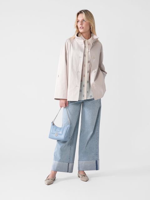 Eine Person steht vor einem neutralen Hintergrund. Sie trägt eine beige Jacke, ein gemustertes Hemd und weite Jeans. Sie hält eine hellblaue Handtasche in der Hand und trägt beige Schuhe. Der Blick ist leicht zur Seite gerichtet und der Ausdruck ist ruhig.