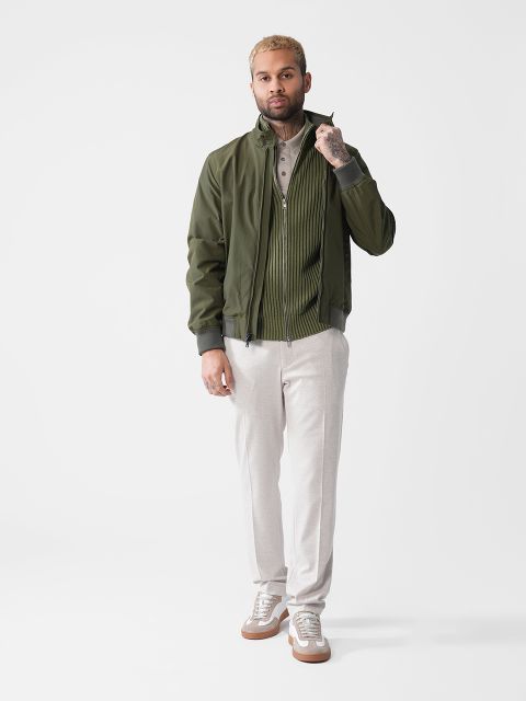 Ein Mann mit kurzen Haaren und Bart steht selbstbewusst vor einem schlichten Hintergrund. Er trägt eine grüne Jacke über einem gestreiften Hemd und einer hellen Hose sowie Turnschuhen. Seine linke Hand ist vor seinem Gesicht erhoben und zeigt einen tätowierten Arm.