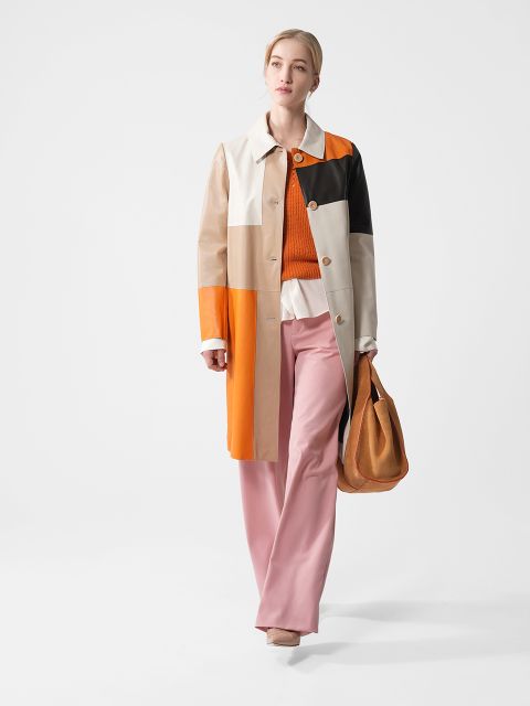 Eine Person in einem bunten Patchworkmantel, einem orangefarbenen Pullover, einer weißen Bluse und einer rosa Hose geht mit einer hellbraunen Tasche in der Hand. Sie hat helles Haar zurückgebunden und trägt beige Schuhe vor einem schlichten Hintergrund.