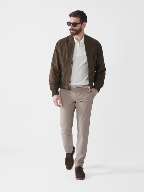 Ein Mann mit Sonnenbrille, einer dunkelbraunen Jacke, einem weißen Hemd, beigen Hosen und dunklen Slippern geht selbstbewusst vor einem schlichten weißen Hintergrund.