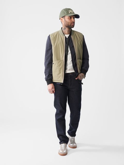 Mann mit grüner Baseballkappe, zweifarbiger Jacke, weißem Hemd, dunklen Jeans und Turnschuhen steht vor einem schlichten Hintergrund.