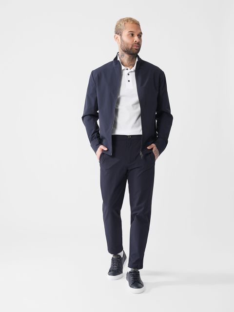 Eine Person mit kurzen Haaren und Bart trägt eine dunkle Jacke, ein weißes Hemd, eine dunkle Hose und dunkle Turnschuhe. Sie steht vor einem schlichten weißen Hintergrund und blickt zur Seite.
