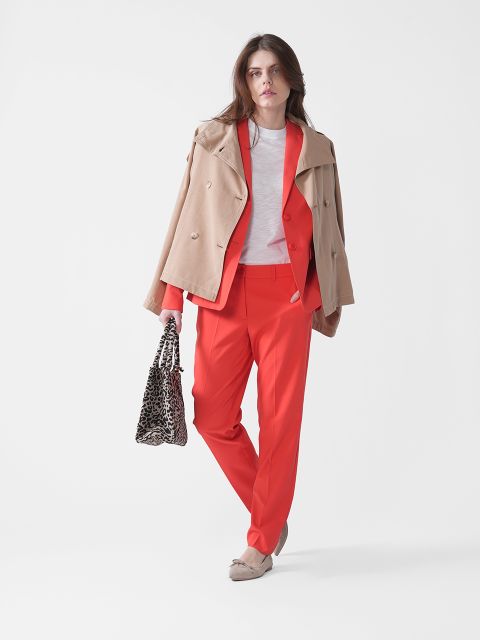 Frau in stilvollem Outfit mit beigem Mantel über einem roten Anzug, weißem Hemd und einer Tasche mit Leopardenmuster in der Hand. Sie steht selbstbewusst vor einem weißen Hintergrund und trägt beige Slipper.