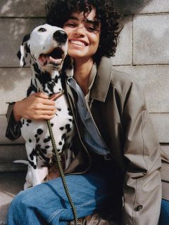 Eine Person mit lockigem Haar, bekleidet mit einem beigen Mantel und blauen Jeans, sitzt lächelnd auf Betonstufen. Sie umarmt einen Dalmatiner, der hechelt und in die Kamera blickt. Der Hund hat eine grüne Leine.
