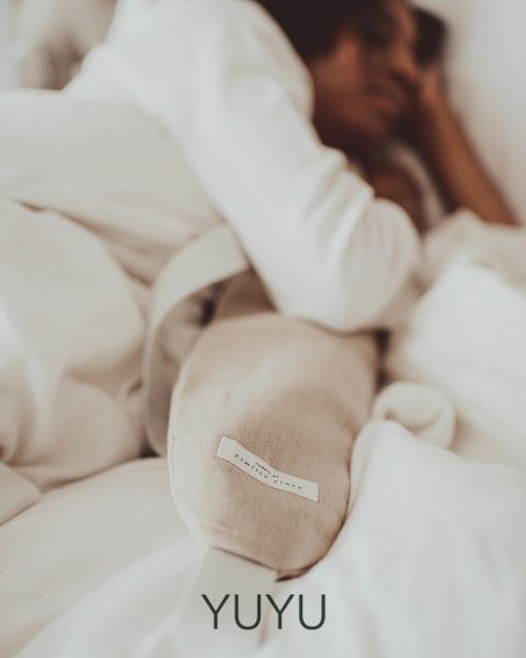 Eine Person liegt in einem Bett, eingehüllt in eine weiße Decke, und hält ein langes, helles Kissen oder Heizkissen in der Hand. Die Atmosphäre ist ruhig und behaglich und vermittelt Geborgenheit und Entspannung. Unten steht das Wort „YUYU“.
