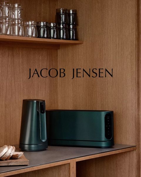 Eine moderne Küche mit Holzregalen, Gläsern, einem Laib Brot und dunkelgrünen Jacob Jensen-Geräten auf dem Tresen. Der Markenname "JACOB JENSEN" steht an der Holzpaneelwand.