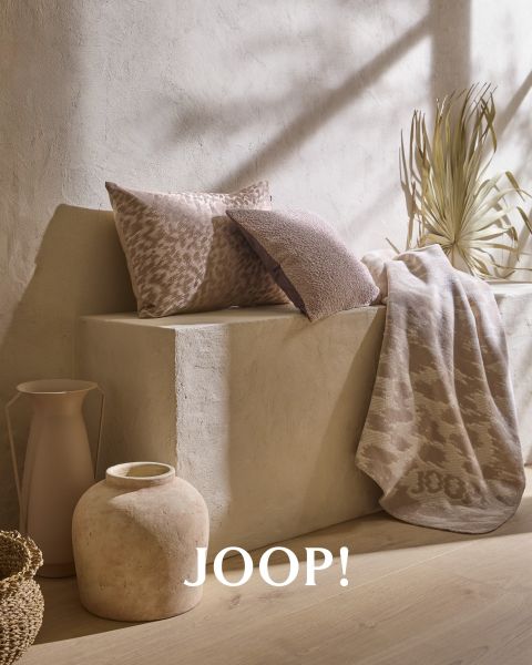 Kissen in neutralen Farben mit Mustern und eine passende Decke sind auf einer minimalistischen Betonbank angeordnet. Daneben stehen eine große Vase und eine dekorative Trockenpflanze. Auf der Decke steht „JOOP!“. Sanftes Sonnenlicht wirft Schatten über die Szene.