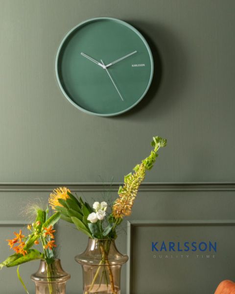 Eine minimalistische grüne Wanduhr mit weißen Zeigern hängt an einer grünen Wand über zwei Glasvasen mit verschiedenen orangefarbenen, weißen und gelben Blumen. In der unteren rechten Ecke erscheint in Blau das Wort „KARLSSON“.