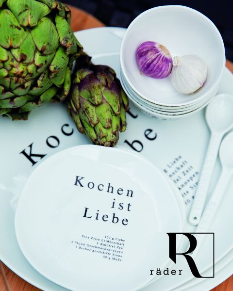 Weiße Teller mit dem deutschen Text „Kochen ist Liebe“ werden zusammen mit Artischocken und Schalen mit Knoblauch ausgestellt. Das Arrangement umfasst weiße Löffel und das Logo „Räder“ auf einem Teller.