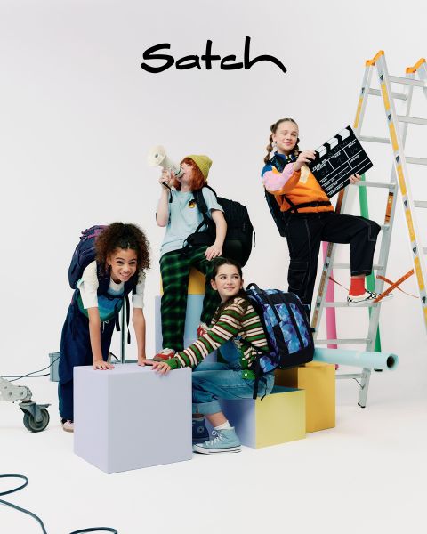Vier Kinder mit Rucksäcken posieren kreativ in einem Studio. Eines steht auf einer Leiter und hält eine Filmklappe, ein anderes spricht in ein Megafon, zwei weitere knien und sitzen auf bunten Würfeln. Studioequipment ist zu sehen.