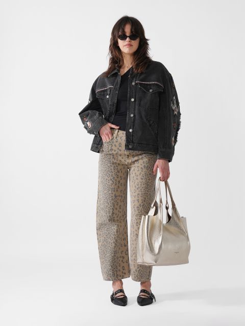 Eine Frau mit Sonnenbrille trägt eine schwarze Jeansjacke und eine Hose mit Leopardenmuster. Sie hält eine metallische Tasche in der Hand und steht vor einem weißen Hintergrund und blickt selbstbewusst in die Kamera.