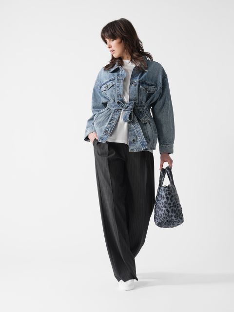 Eine Person steht vor einem weißen Hintergrund und trägt eine Jeansjacke mit Gürtel über einem weißen Hemd, dazu eine schwarze Nadelstreifenhose und weiße Turnschuhe. Sie trägt eine blau gemusterte Handtasche.