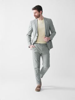Mann trägt einen hellgrauen Anzug, einen cremefarbenen Pullover und braune Slipper. Er hat eine Hand in der Tasche und blickt beim Gehen zur Seite. Der Hintergrund ist schlicht weiß.