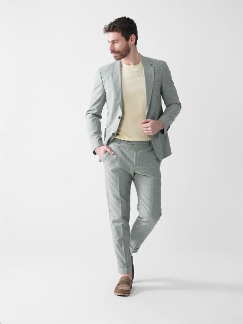 Mann trägt einen hellgrauen Anzug, einen cremefarbenen Pullover und braune Slipper. Er hat eine Hand in der Tasche und blickt beim Gehen zur Seite. Der Hintergrund ist schlicht weiß.