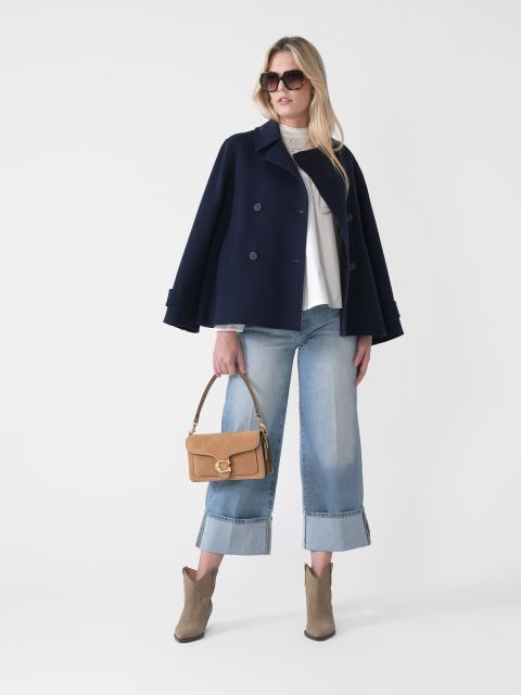 Eine Frau steht vor einem schlichten Hintergrund. Sie trägt einen marineblauen Mantel, ein weißes Oberteil und hellblaue Jeans mit weitem Bein. Sie hält eine hellbraune Handtasche in der Hand, trägt eine Sonnenbrille und hellbraune Stiefeletten. Ihre Hände sind in die Hüften gestemmt, und ihr langes Haar ist offen.