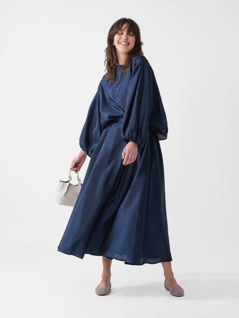 Eine Person mit langen braunen Haaren lächelt und trägt ein fließendes dunkelblaues Kleid mit bauschigen Ärmeln. Sie hält eine kleine weiße Handtasche in der Hand und steht vor einem schlichten weißen Hintergrund.