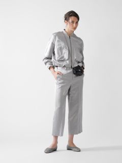 Eine Person in glänzender silberner Jacke und passender Hose posiert vor einem schlichten Hintergrund. Sie hat eine Hand in der Tasche und trägt eine schwarze Gürteltasche. Ihre Haare sind hochgesteckt, und sie trägt silberne Ballerinas.