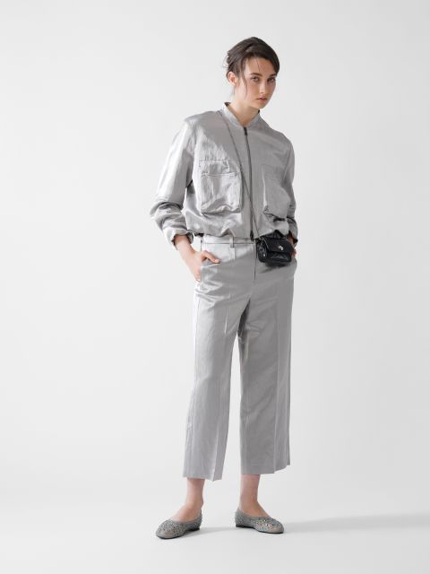 Eine Person in glänzender silberner Jacke und passender Hose posiert vor einem schlichten Hintergrund. Sie hat eine Hand in der Tasche und trägt eine schwarze Gürteltasche. Ihre Haare sind hochgesteckt, und sie trägt silberne Ballerinas.