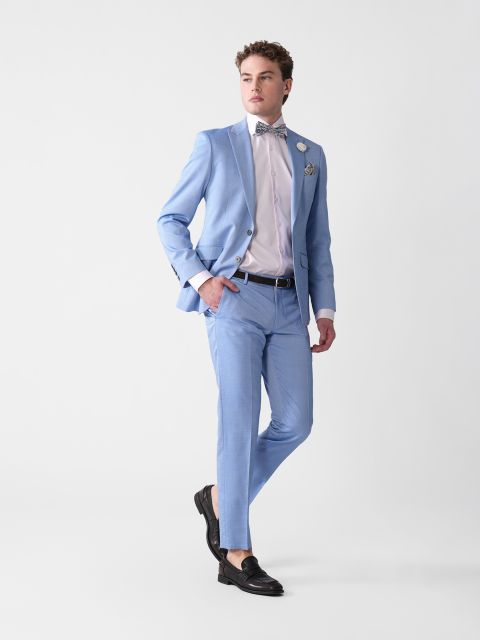 Eine Person in einem hellblauen Anzug mit weißem Hemd und gemusterter Fliege posiert vor einem schlichten Hintergrund. Sie trägt schwarze Slipper und hat als Accessoires ein Einstecktuch und eine Blume am Revers. Eine Hand steckt in der Hosentasche, der Blick ist zur Seite gerichtet.