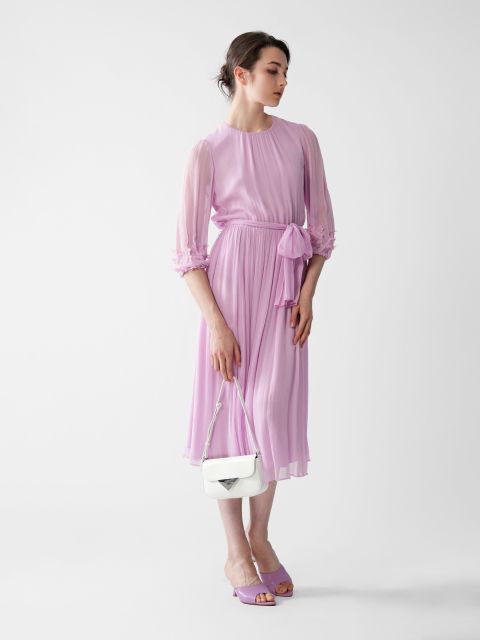 Eine Frau steht anmutig da und trägt ein fließendes lavendelfarbenes Kleid mit transparenten Ärmeln und einer passenden Taillenschärpe. Sie hält eine kleine weiße Handtasche in der Hand und trägt lila Sandalen. Der schlichte weiße Hintergrund unterstreicht die Eleganz ihres Outfits.