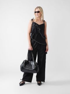 Eine Frau mit langen blonden Haaren trägt ein ärmelloses schwarzes Top mit Rüschen, eine schwarze Hose und spitze Schuhe. Sie hält eine große schwarze Handtasche in der Hand und trägt eine Sonnenbrille. Der Hintergrund ist schlicht weiß.