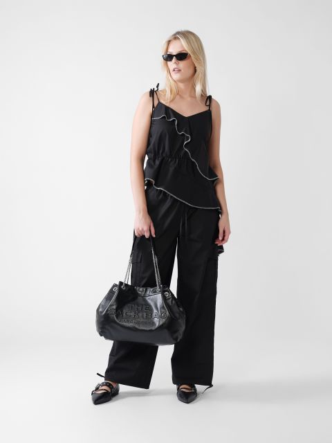 Eine Frau mit langen blonden Haaren trägt ein ärmelloses schwarzes Top mit Rüschen, eine schwarze Hose und spitze Schuhe. Sie hält eine große schwarze Handtasche in der Hand und trägt eine Sonnenbrille. Der Hintergrund ist schlicht weiß.