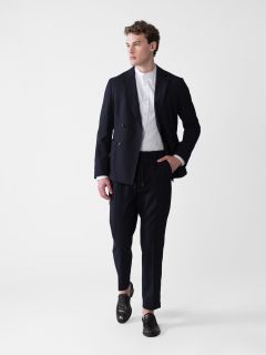 Eine Person in dunklem Anzug und weißem Hemd steht vor einem schlichten Hintergrund. Sie hat eine Hand in der Tasche und blickt zur Seite. Dunkle Schuhe runden das Outfit ab.