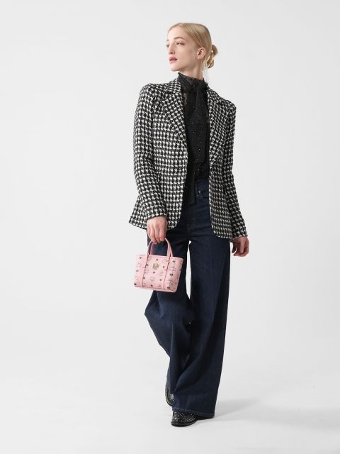 Eine Person mit blondem Haar im Dutt trägt einen schwarz-weißen Blazer mit Hahnentrittmuster, ein schwarzes Hemd und weite Jeans. Sie hält eine kleine rosa Handtasche in der Hand und steht vor einem schlichten weißen Hintergrund.