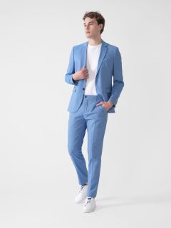 Eine Person in einem hellblauen Anzug, einem weißen T-Shirt und weißen Turnschuhen steht vor einem schlichten weißen Hintergrund. Sie hat eine Hand in der Tasche, die andere berührt das Revers ihrer Jacke und blickt zur Seite.
