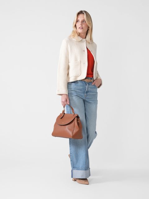 Eine Frau in einer weißen Strukturjacke und einem roten Oberteil, in weiten Jeans und beigen Pumps, hält eine braune Lederhandtasche in der Hand und steht vor einem schlichten weißen Hintergrund.