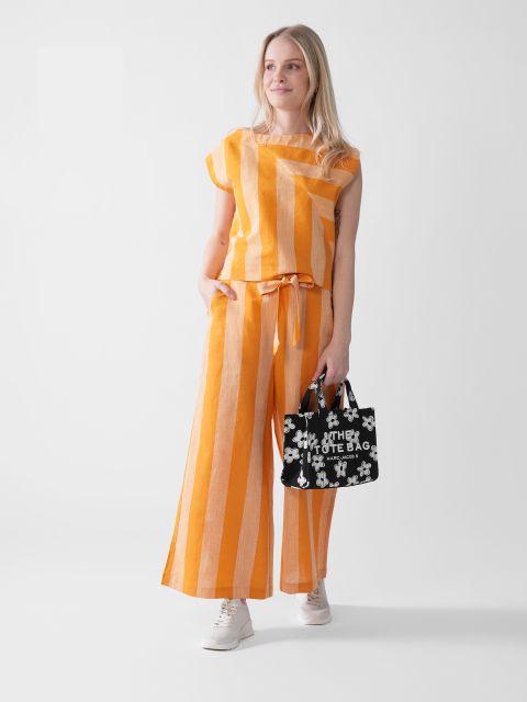 Eine Person in einem orange-weiß gestreiften Outfit und einer schwarz-weißen Einkaufstasche steht vor einem schlichten Hintergrund. Sie lächelt und trägt weiße Turnschuhe.