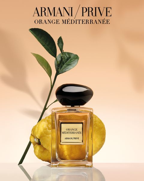 Vor einer gelben Zitrusfrucht und einem grünen, belaubten Zweig steht ein Flakon des Parfüms „Armani/Prive Orange Méditerranée“ vor einem zarten pfirsichfarbenen Hintergrund.