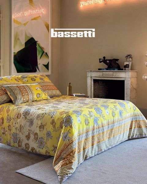 Ein Schlafzimmer mit einer gelb geblümten Tagesdecke, passenden Kissen, einem weißen Marmorkamin, moderner Kunst an der Wand und einer Neonreklame darüber mit der Aufschrift „Gestern, heute“. Das Wort „Bassetti“ ist deutlich zu sehen.