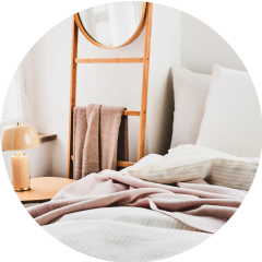 Eine gemütliche Schlafzimmerecke mit einem Bett mit weißer und rosa Bettwäsche. An der Wand lehnt eine Holzleiter mit einem runden Spiegel und Handtüchern. Auf einem hölzernen Nachttisch steht eine kleine beige Lampe. Natürliches Licht fällt durch ein nahes Fenster.