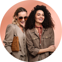 Zwei stilvolle Frauen lächeln vor einem pfirsichfarbenen Hintergrund. Die eine trägt eine Sonnenbrille, ein kariertes Kostüm und eine braune Clutch; die andere trägt eine braune Jacke mit Schal und hat lockiges Haar.