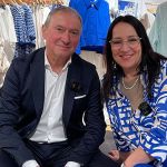 Ein Mann und eine Frau sitzen lächelnd in einem Bekleidungsgeschäft. Sie tragen aufeinander abgestimmte blau-weiße Outfits. Hinter ihnen stehen Kleiderständer mit blau-weiß-roter Kleidung.