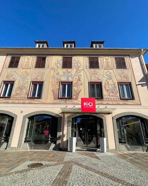 Historisches Gebäude mit kunstvollen Fresken über Bogenfenstern und -türen. Über dem Eingang steht ein rot-weißes Schild mit der Aufschrift „K&U“. Die Fassade besticht durch kunstvolle Muster und einen strahlend blauen Himmel im Hintergrund.