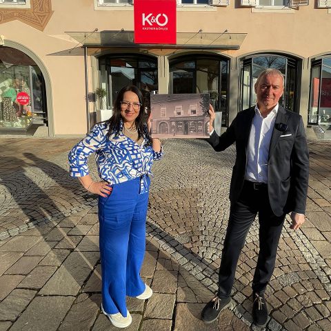 Zwei Personen stehen vor einem Gebäude mit einem „K&O Kastner & Öhler“-Schild. Die Frau trägt blau-weiße Kleidung, der Mann einen schwarzen Anzug. Sie halten ein altes Schwarz-Weiß-Foto einer ähnlichen Szene in den Händen. Im Vordergrund ist Kopfsteinpflaster zu sehen.