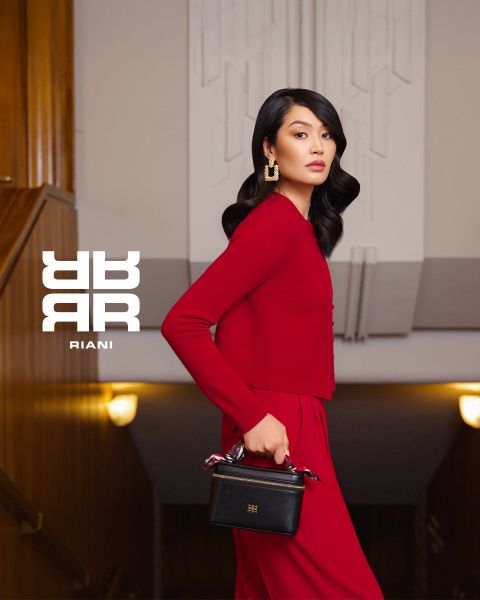 Eine Frau mit langen dunklen Haaren, die ein rotes Kleid und goldene Ohrringe trägt, steht in einem Haus und hält eine schwarze Handtasche. Sie posiert auf einer Treppe mit Holzvertäfelung und Art-déco-Beleuchtung. Neben ihr ist das RIANI-Logo zu sehen.